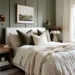 Modern bedroom with an olive-green paneled wall, matching green pillows, cream bedding, and brass wall lights