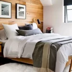 Small modern bedroom with warm wood walls, wall-mounted lights, simple furniture, and white bedding