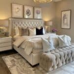 cream bedroom with cream and white bedding, tan white and beige throw pillows, bench lavender and cream wall art, chandelier ceiling light table lamps
