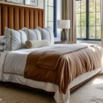 Bedroom with a tall camel channel-tufted headboard, layered white bedding, a warm brown duvet, neutral curtains, and black-framed windows.