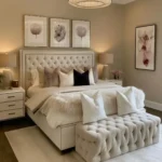 Light neutral cozy luxury bedroom with a tufted cream bed, layered bedding, upholstered bench, crystal chandelier, and soft area rug.