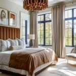 Luxury bedroom with a camel channel-tufted headboard, layered cream and rust bedding, black-framed windows, floor-to-ceiling curtains, and a brass chandelier