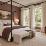 Calm bedroom with muted plum walls, a black canopy bed, layered cream bedding, a brown reading chair, and tall windows overlooking leafy trees.