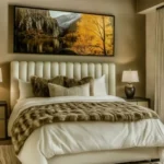 Neutral bedroom with a cream upholstered bed, layered white and brown bedding, matching bedside lamps, and large landscape art above the headboard.