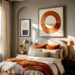 Warm neutral bedroom with gray carpet, a curved cream upholstered bed, rust-colored pillows and throw, brass lamps, and large circular wall art.